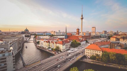 Die Aussicht zeigt die Stadt Berlin mit der Spree, umgeben von historischen Gebäuden und modernen Wolkenkratzern. Der Fernsehturm ist im Hintergrund sichtbar. Im Vordergrund verläuft eine belebte Straße, die über eine Brücke führt, während weitere Stadtteile im Hintergrund sanft in der Dämmerung leuchten.