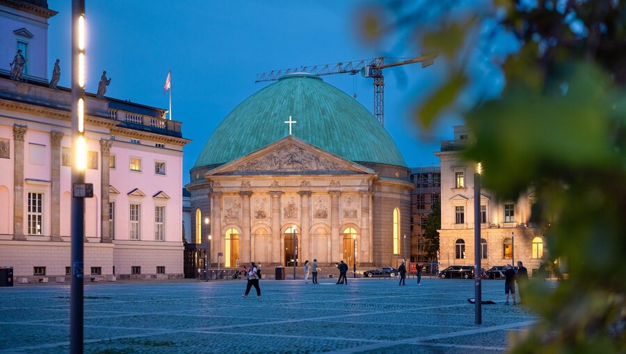 Die Bildbeschreibung zeigt einen historischen Bau mit einer grünen Kuppel, gekrönt von einem Kreuz. Der Eingang wird von warmem Licht beleuchtet. Im Vordergrund sind Menschen auf einem gepflasterten Platz zu sehen, umgeben von Gebäuden und Straßenlaternen in der Dämmerung.