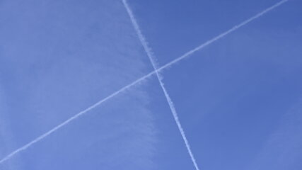 Der Himmel ist klar und blau, durchzogen von mehreren weißen Kondensstreifen, die sich kreuzen und ein Muster bilden. Die Linien verlaufen in diagonalen Richtungen und verleihen dem Himmel ein strukturiertes Aussehen.