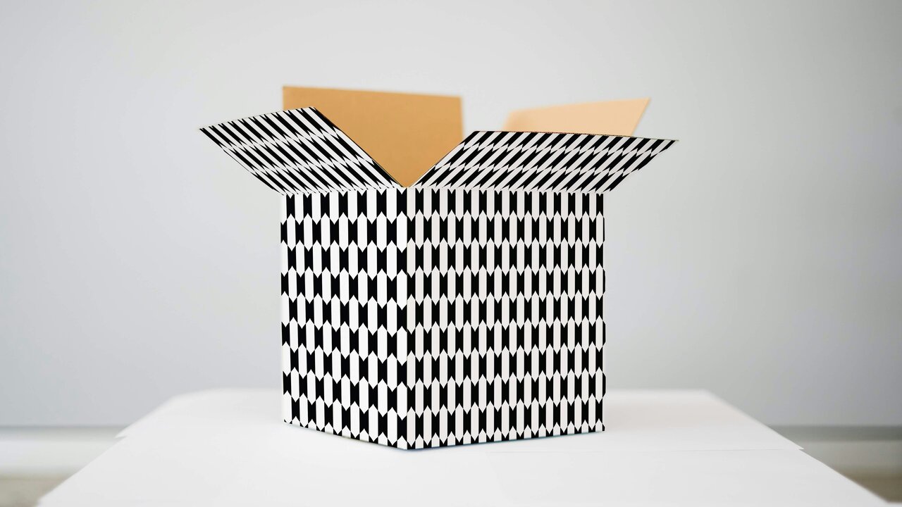 Eine offene Kartonschachtel mit einem geometrischen Muster in Schwarz und Weiß steht auf einem Tisch. Die Schachtel hat einen quadratischen Boden und zeigt ihre Innenseite. Der Hintergrund ist neutral und unscharf.