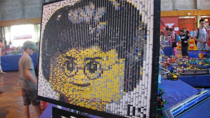 Das Bild zeigt eine große Mosaik-Art aus Lego-Steinen, die das Gesicht eines Lego-Charakters darstellt. Die Farben des Mosaiks sind überwiegend Gelb für das Gesicht und Schwarz für das Haar. Es ist Teil einer Ausstellung mit verschiedenen Lego-Modellen.