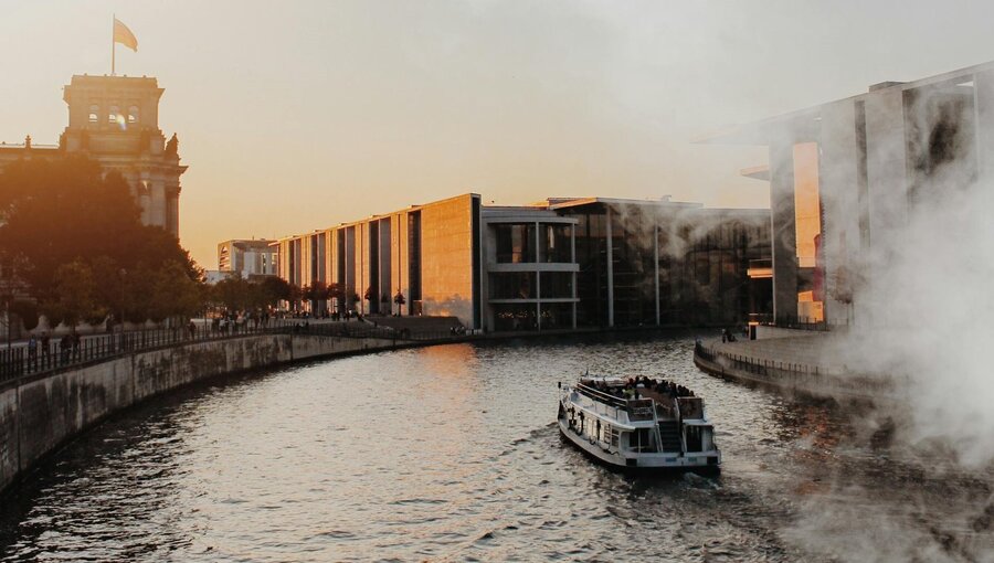 Ein Boot fährt auf einem Fluss, umgeben von Gebäuden. Die Sonne steht tief am Horizont und taucht die Szene in warme Farben. Der Dampf im Hintergrund verstärkt die entspannte Atmosphäre des Abends in einer städtischen Umgebung.