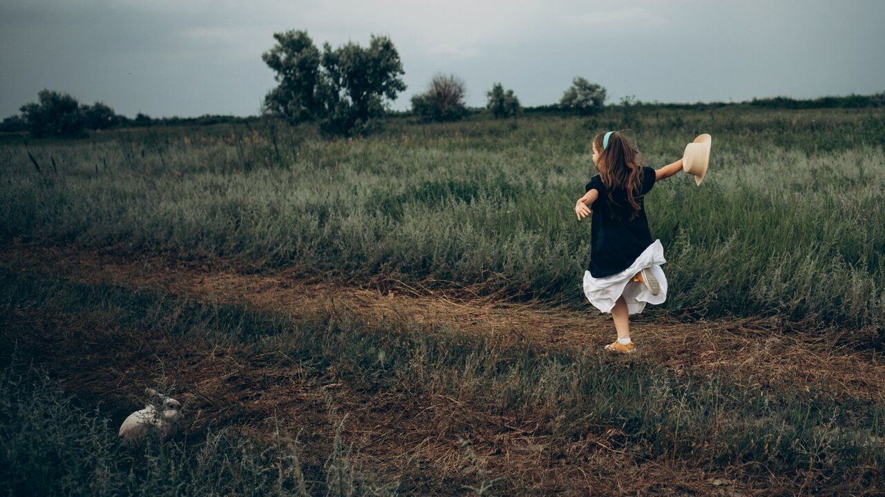 EinKind mit langen Haaren trägt ein schwarzes Oberteil und einen weißen Rock. Es läuft fröhlich auf einem schmalen Weg durch ein grünes Feld, während es einen Hut in der Hand hält. Im Hintergrund sind vereinzelte Bäume und ein bewölkter Himmel zu sehen.