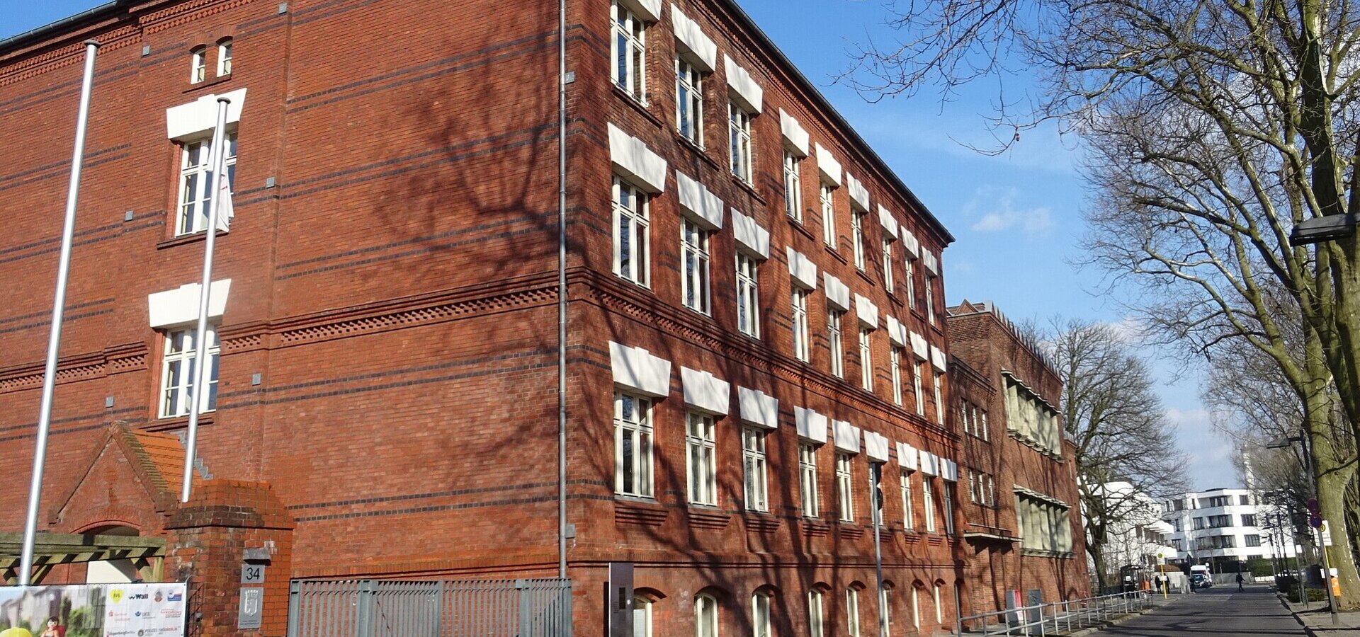Das Bild zeigt ein rotes Ziegelgebäude im Vordergrund, das eine auffällige Architektur mit mehreren Fenstern hat. Im Hintergrund sind Bäume und ein Teil einer Straße zu sehen, der eine ruhige städtische Umgebung vermittelt. Es ist die Thalia-Grundschule in Berlin.