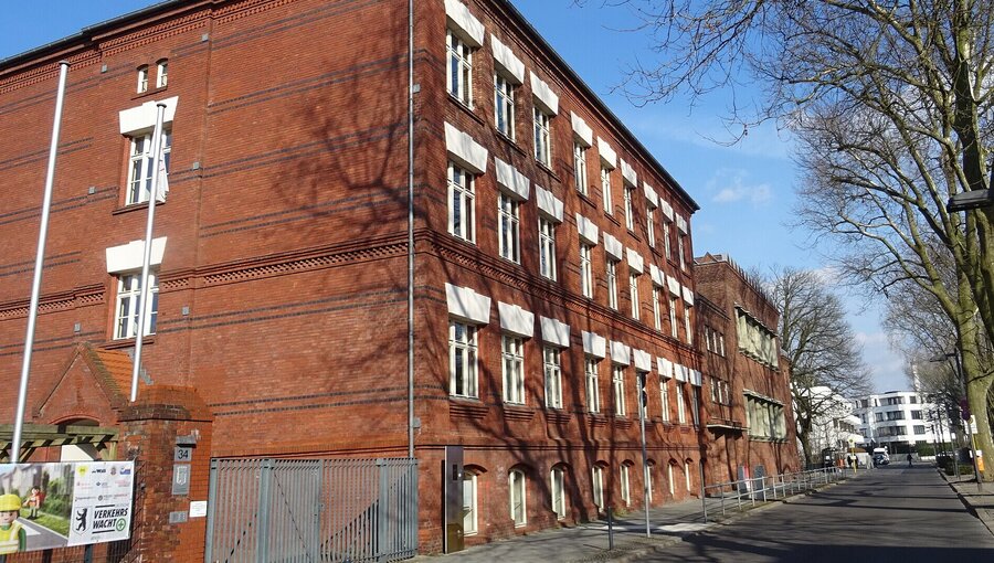 Das Bild zeigt ein rotes Ziegelgebäude im Vordergrund, das eine auffällige Architektur mit mehreren Fenstern hat. Im Hintergrund sind Bäume und ein Teil einer Straße zu sehen, der eine ruhige städtische Umgebung vermittelt. Es ist die Thalia-Grundschule in Berlin.