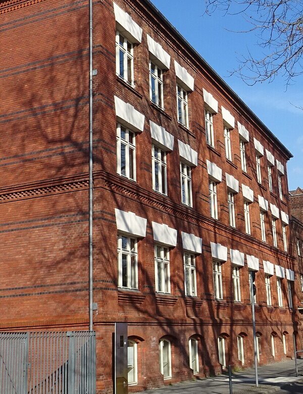Das Bild zeigt ein rotes Ziegelgebäude im Vordergrund, das eine auffällige Architektur mit mehreren Fenstern hat. Im Hintergrund sind Bäume und ein Teil einer Straße zu sehen, der eine ruhige städtische Umgebung vermittelt. Es ist die Thalia-Grundschule in Berlin.