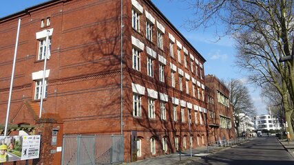 Das Bild zeigt ein rotes Ziegelgebäude im Vordergrund, das eine auffällige Architektur mit mehreren Fenstern hat. Im Hintergrund sind Bäume und ein Teil einer Straße zu sehen, der eine ruhige städtische Umgebung vermittelt. Es ist die Thalia-Grundschule in Berlin.