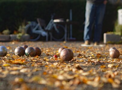 Eine Boule-Bahn ist mit bunten Blättern bedeckt. Mehrere Kugeln in verschiedenen Farben liegen auf dem Boden, während eine Person im Hintergrund steht. Ein Fahrrad ist ebenfalls sichtbar.