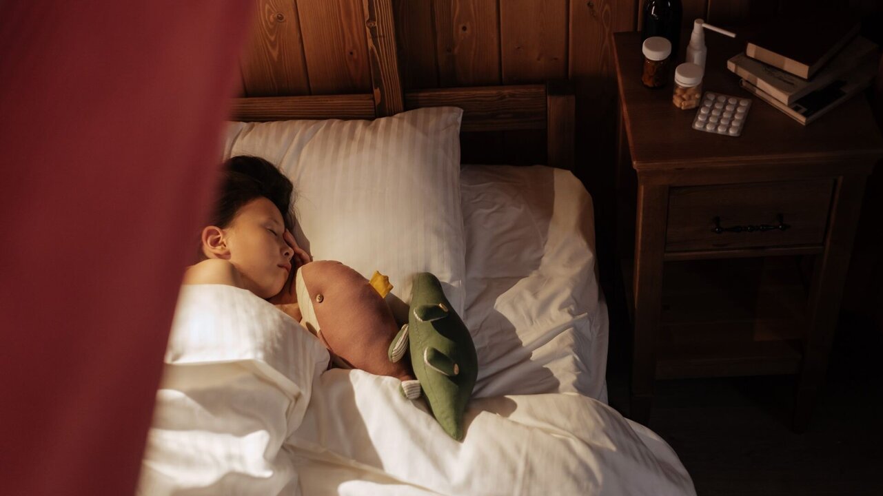 Ein Kind schläft friedlich in einem Bett, umgeben von weißen Bettdecken. Es hält zwei Plüschtiere, eines rosa und das andere grün, in den Armen. Im Hintergrund sind ein Nachttisch mit Flaschen und einer Tablettenpackung sichtbar. Weiche, warme Lichtverhältnisse schaffen eine gemütliche Atmosphäre.