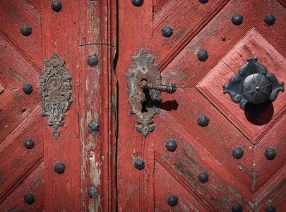 Eine alte rote Holztür mit diagonalen Mustern. Die Tür hat große, schwarze Nägel und dekorative Beschläge aus Metall. Ein Schlüsselhalter und ein runder Türknauf sind sichtbar, die insgesamt einen rustikalen und historischen Eindruck vermitteln.