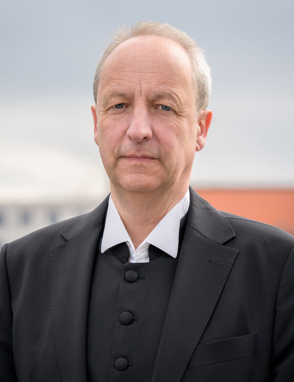 Ein Mann steht im Vordergrund und schaut direkt in die Kamera. Er trägt einen schwarzen Anzug mit einem weißen Hemd. Im Hintergrund sind unscharfe städtische Gebäude und ein Turm sichtbar. Der Himmel ist bewölkt.