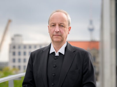 Ein Mann steht im Vordergrund und schaut direkt in die Kamera. Er trägt einen schwarzen Anzug mit einem weißen Hemd. Im Hintergrund sind unscharfe städtische Gebäude und ein Turm sichtbar. Der Himmel ist bewölkt.