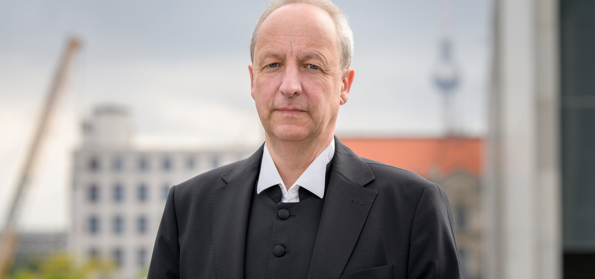 Ein Mann steht im Vordergrund und schaut direkt in die Kamera. Er trägt einen schwarzen Anzug mit einem weißen Hemd. Im Hintergrund sind unscharfe städtische Gebäude und ein Turm sichtbar. Der Himmel ist bewölkt.