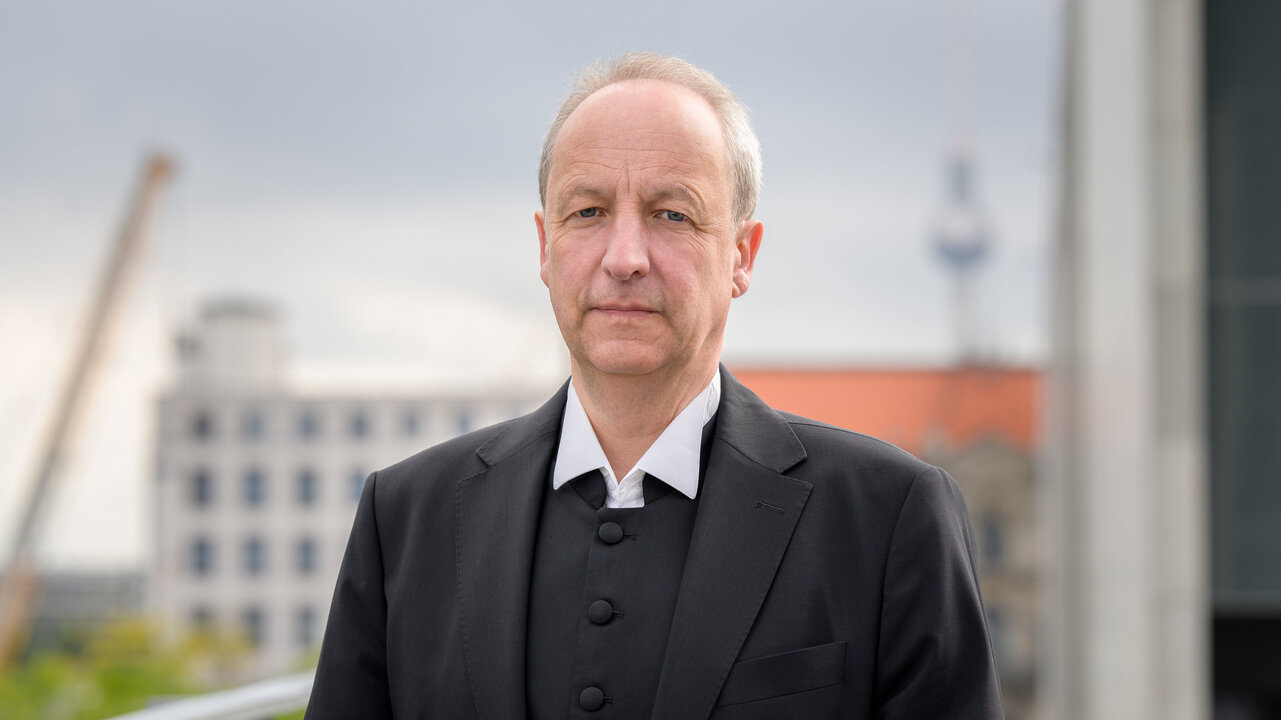 Ein Mann steht im Vordergrund und schaut direkt in die Kamera. Er trägt einen schwarzen Anzug mit einem weißen Hemd. Im Hintergrund sind unscharfe städtische Gebäude und ein Turm sichtbar. Der Himmel ist bewölkt.
