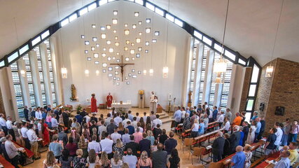 Eine Gruppe von Menschen ist in einer hellen Kirche versammelt. Im Vordergrund stehen mehrere Priester, einer hält eine Ansprache. Im Hintergrund sind kunstvoll angeordnete Fenster und ein Kreuz zu sehen. Die Atmosphäre wirkt feierlich und andächtig.