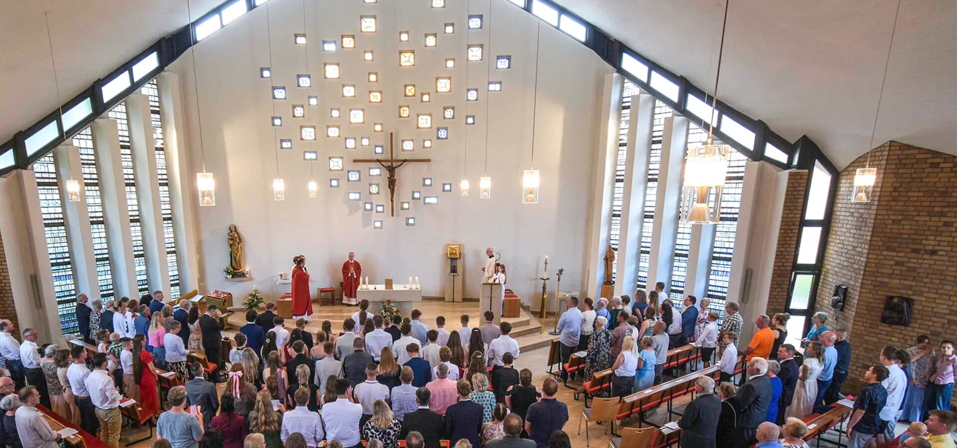 Eine Gruppe von Menschen ist in einer hellen Kirche versammelt. Im Vordergrund stehen mehrere Priester, einer hält eine Ansprache. Im Hintergrund sind kunstvoll angeordnete Fenster und ein Kreuz zu sehen. Die Atmosphäre wirkt feierlich und andächtig.