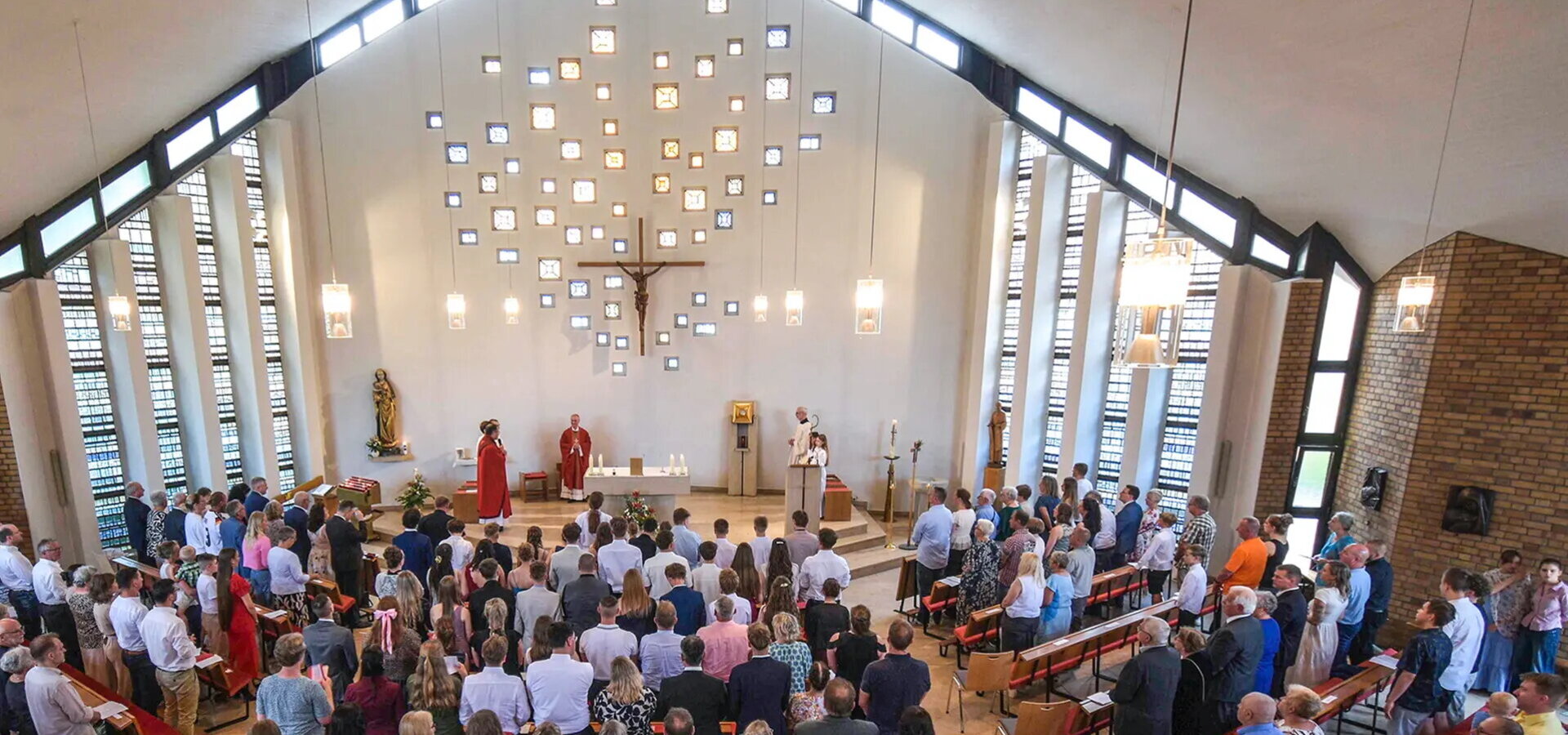 Eine Gruppe von Menschen ist in einer hellen Kirche versammelt. Im Vordergrund stehen mehrere Priester, einer hält eine Ansprache. Im Hintergrund sind kunstvoll angeordnete Fenster und ein Kreuz zu sehen. Die Atmosphäre wirkt feierlich und andächtig.