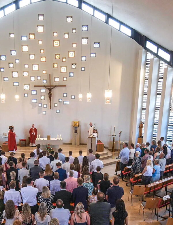 Eine Gruppe von Menschen ist in einer hellen Kirche versammelt. Im Vordergrund stehen mehrere Priester, einer hält eine Ansprache. Im Hintergrund sind kunstvoll angeordnete Fenster und ein Kreuz zu sehen. Die Atmosphäre wirkt feierlich und andächtig.