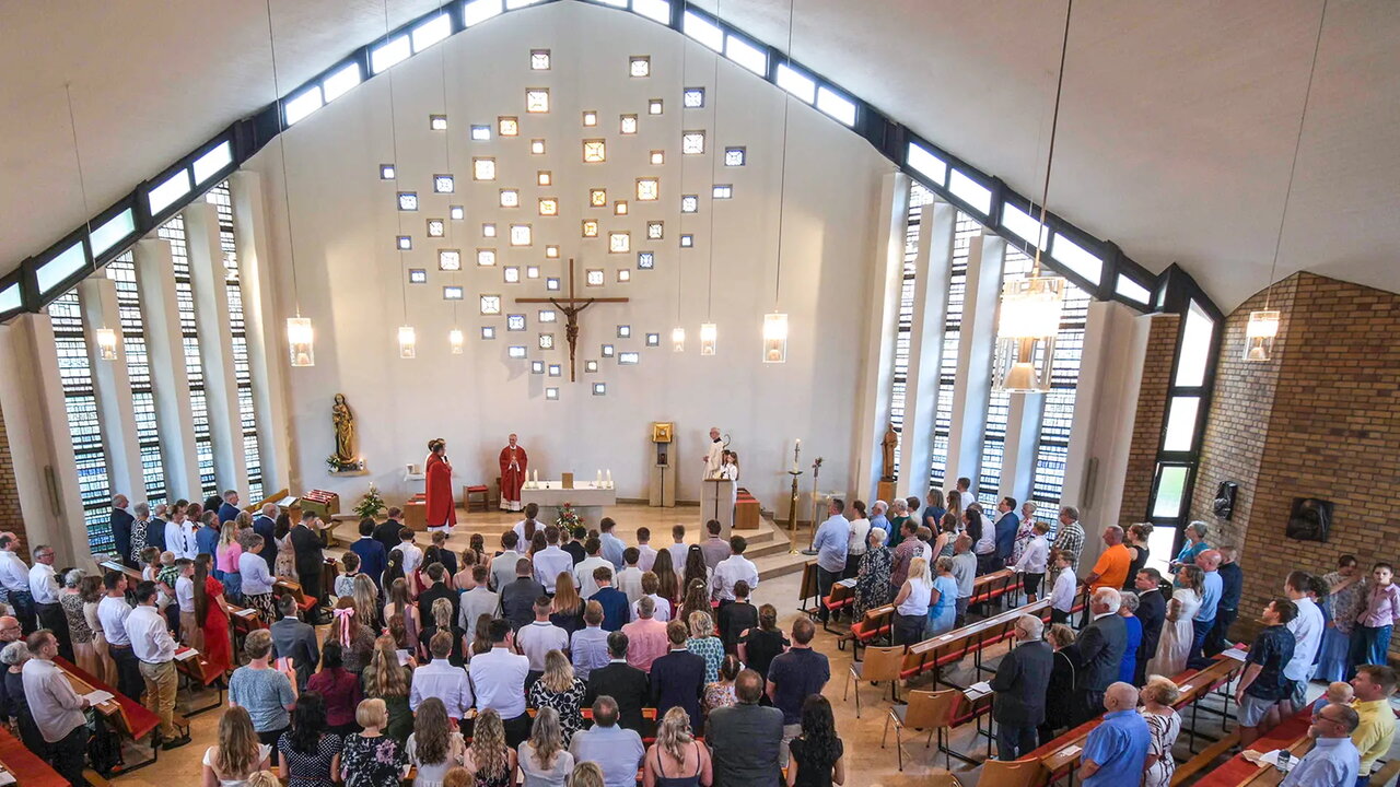 Eine Gruppe von Menschen ist in einer hellen Kirche versammelt. Im Vordergrund stehen mehrere Priester, einer hält eine Ansprache. Im Hintergrund sind kunstvoll angeordnete Fenster und ein Kreuz zu sehen. Die Atmosphäre wirkt feierlich und andächtig.