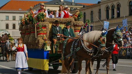 Eine festlich dekorierte Pferdekutsche zieht durch die Straße. Auf der Kutsche sitzen fröhliche Menschen in traditioneller Tracht, umgeben von Blumen und Stroh. Eine Frau in einem roten und weißen traditionellen Kleid geht neben der Kutsche. Die Atmosphäre ist festlich und lebhaft.