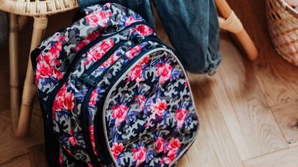 Ein Rucksack mit buntem Blumenmuster liegt auf dem Boden. Daneben befindet sich eine Jeans. Der Hintergrund zeigt einen Stuhl und einen Korb, was eine gemütliche Räumlichkeit vermittelt.