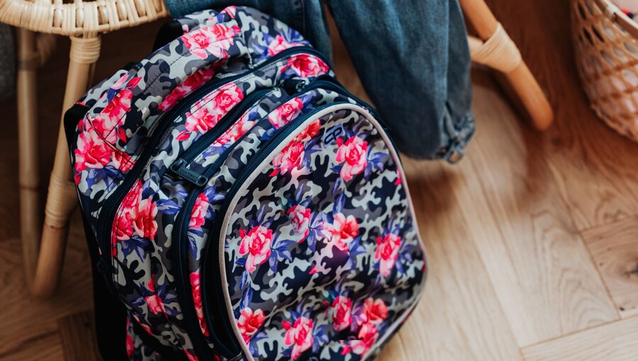 Ein Rucksack mit buntem Blumenmuster liegt auf dem Boden. Daneben befindet sich eine Jeans. Der Hintergrund zeigt einen Stuhl und einen Korb, was eine gemütliche Räumlichkeit vermittelt.