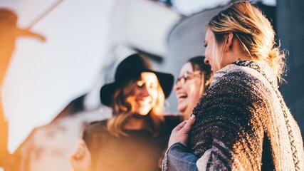 Drei Frauen stehen nah beieinander und lachen. Eine trägt einen Hut und die andere eine Decke über den Schultern. Die Atmosphäre wirkt freundlich und entspannt, möglicherweise bei einem Treffen im Freien während des Sonnenuntergangs.