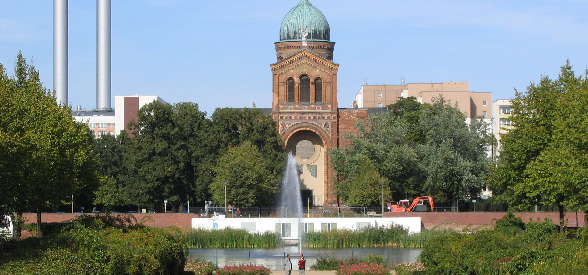 Das Bild zeigt ein historisches Gebäude mit einer markanten Kuppel, umgeben von Bäumen und einem Wasserbereich. Im Vordergrund schwimmt ein Schwan auf dem See, während sich auf der Terrasse des Gebäudes Menschen versammelt haben. Der Himmel ist klar und blau.