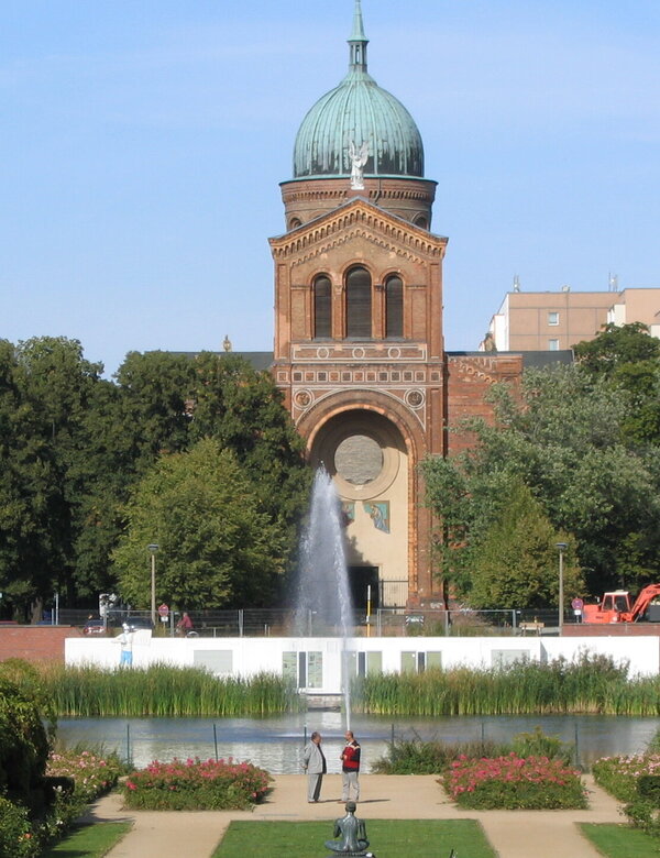Das Bild zeigt ein historisches Gebäude mit einer markanten Kuppel, umgeben von Bäumen und einem Wasserbereich. Im Vordergrund schwimmt ein Schwan auf dem See, während sich auf der Terrasse des Gebäudes Menschen versammelt haben. Der Himmel ist klar und blau.