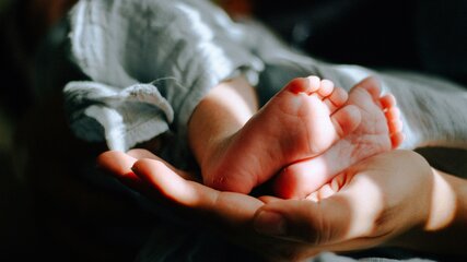 Eine Hand hält die Füße eines Neugeborenen. Die kleinen, zarten Füße sind deutlich zu sehen, während der Hintergrund unscharf bleibt. Die Szene vermittelt ein Gefühl von Wärme und Geborgenheit.