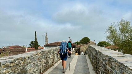 Zwei Wanderer gehen auf einer steinernen Brücke, umgeben von historischen Gebäuden und grünen Bäumen. Die Atmosphäre ist ruhig, mit einem bewölkten Himmel im Hintergrund. Sie tragen Rucksäcke und nutzen Wanderstöcke.