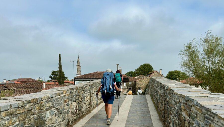 Zwei Wanderer gehen auf einer steinernen Brücke, umgeben von historischen Gebäuden und grünen Bäumen. Die Atmosphäre ist ruhig, mit einem bewölkten Himmel im Hintergrund. Sie tragen Rucksäcke und nutzen Wanderstöcke.