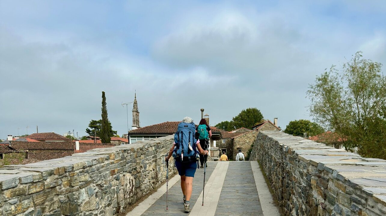 Zwei Wanderer gehen auf einer steinernen Brücke, umgeben von historischen Gebäuden und grünen Bäumen. Die Atmosphäre ist ruhig, mit einem bewölkten Himmel im Hintergrund. Sie tragen Rucksäcke und nutzen Wanderstöcke.