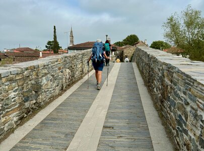Zwei Wanderer gehen auf einer steinernen Brücke, umgeben von historischen Gebäuden und grünen Bäumen. Die Atmosphäre ist ruhig, mit einem bewölkten Himmel im Hintergrund. Sie tragen Rucksäcke und nutzen Wanderstöcke.