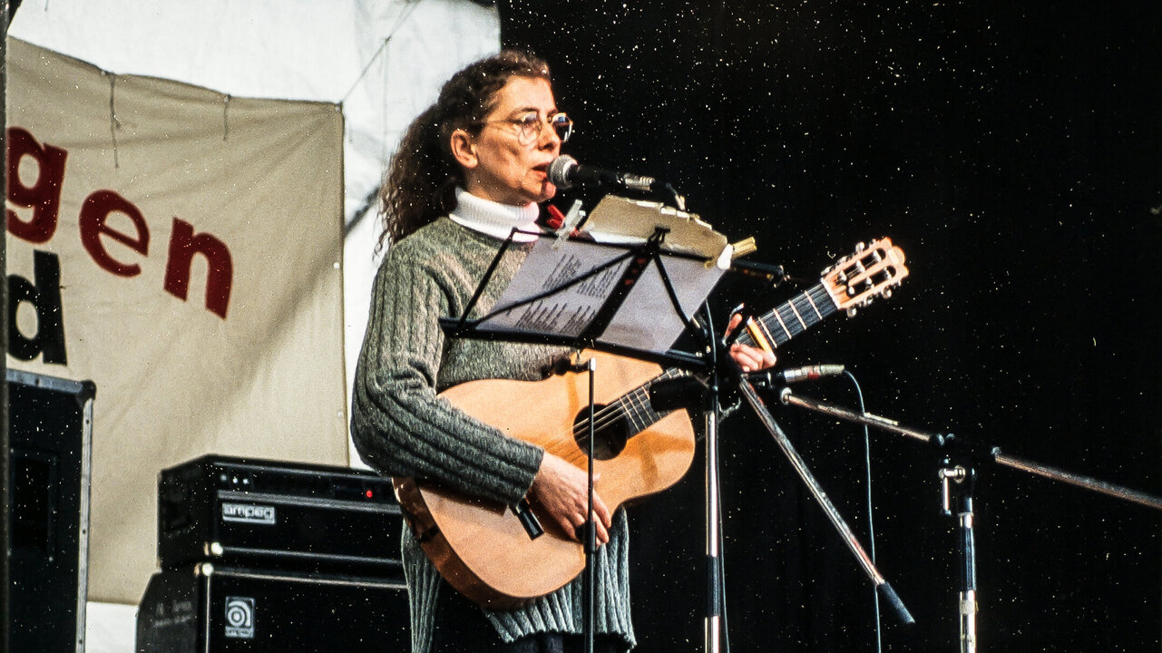 Eine Frau mit lockigem Haar steht auf einer Bühne und spielt Gitarre. Sie trägt einen grauen Pullover und gestreifte Strumpfhosen. Ihre Mimik zeigt Leidenschaft für die Musik, während sie ein Mikrofon benutzt. Im Hintergrund sind Lautsprecher und eine musikalische Umgebung sichtbar.