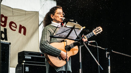 Eine Frau mit lockigem Haar steht auf einer Bühne und spielt Gitarre. Sie trägt einen grauen Pullover und gestreifte Strumpfhosen. Ihre Mimik zeigt Leidenschaft für die Musik, während sie ein Mikrofon benutzt. Im Hintergrund sind Lautsprecher und eine musikalische Umgebung sichtbar.