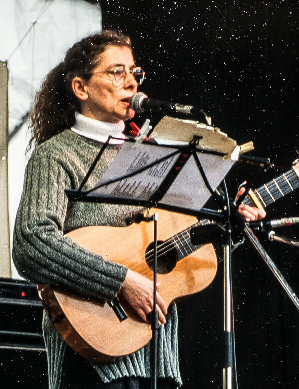 Eine Frau mit lockigem Haar steht auf einer Bühne und spielt Gitarre. Sie trägt einen grauen Pullover und gestreifte Strumpfhosen. Ihre Mimik zeigt Leidenschaft für die Musik, während sie ein Mikrofon benutzt. Im Hintergrund sind Lautsprecher und eine musikalische Umgebung sichtbar.