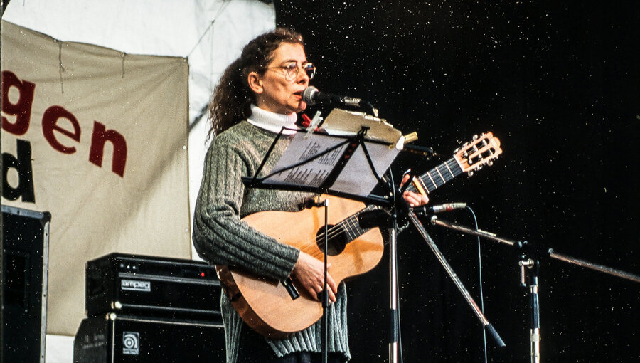 Eine Frau mit lockigem Haar steht auf einer Bühne und spielt Gitarre. Sie trägt einen grauen Pullover und gestreifte Strumpfhosen. Ihre Mimik zeigt Leidenschaft für die Musik, während sie ein Mikrofon benutzt. Im Hintergrund sind Lautsprecher und eine musikalische Umgebung sichtbar.