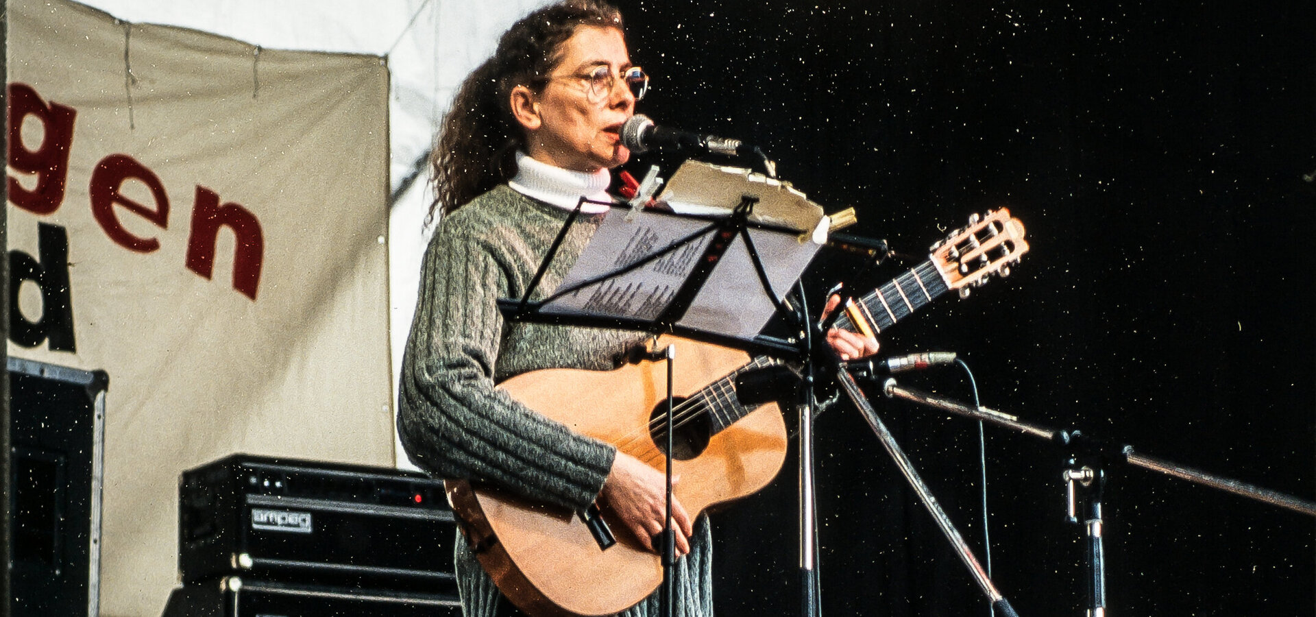 Eine Frau mit lockigem Haar steht auf einer Bühne und spielt Gitarre. Sie trägt einen grauen Pullover und gestreifte Strumpfhosen. Ihre Mimik zeigt Leidenschaft für die Musik, während sie ein Mikrofon benutzt. Im Hintergrund sind Lautsprecher und eine musikalische Umgebung sichtbar.
