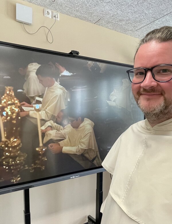 Ein Mann in einem hellen Gewand steht vor einem Bildschirm, auf dem eine Szene mit Kerzen und Menschen, die sich um einen Tisch versammeln, zu sehen ist. Der Mann lächelt und trägt eine Brille. Im Hintergrund sind einige Unterrichtsmaterialien sichtbar.