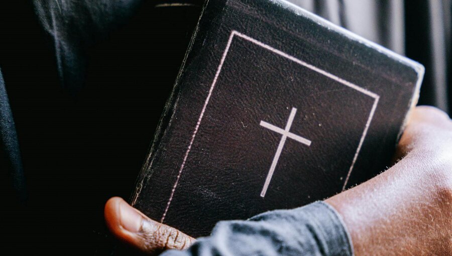 Eine Hand hält eine schwarze Bibel mit einem weißen Kreuz auf dem Cover. Der Hintergrund ist unscharf und dunkel, was die Bibel in den Vordergrund rückt. Die Hand zeigt eine enge Verbindung zum religiösen Text.