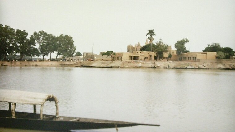 Ein ruhiger Fluss mit einem Holzboot im Vordergrund. Am Ufer sind einige Bäume und eine Baugruppe sichtbar, die aus traditionellen, erdigen Strukturen besteht. Im Hintergrund ragt ein Tempelgebäude gegen den Himmel.