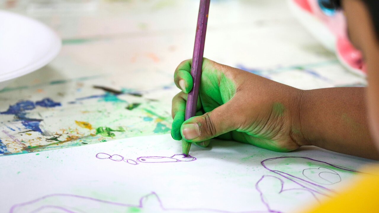 Ein Kind hält einen bunten Stift in der Hand und zeichnet auf ein Blatt Papier. Die Hand ist grün gefärbt, offensichtlich vom Malen. Der Tisch ist mit Farbspritzern und weiteren Malutensilien bedeckt.