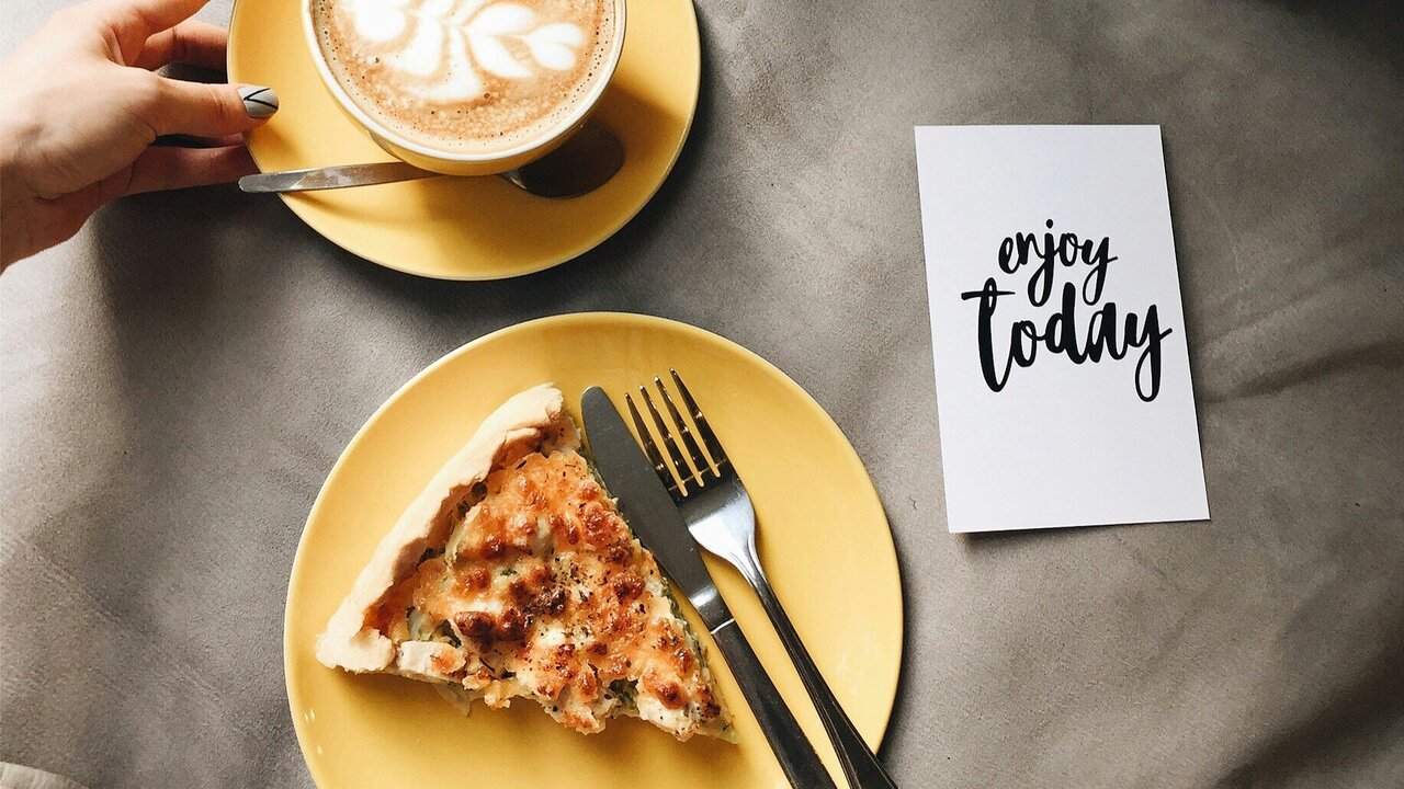 Eine Hand hält eine Tasse mit Kaffee, auf dem ein Herzmuster zu sehen ist. Daneben liegt ein gelbes Teller mit einem Stück Pizza und Besteck. Auf einem weißen Kartenblatt steht in schöner Schrift "enjoy today".