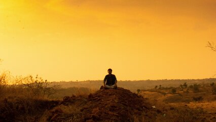 Eine Person sitzt auf einem kleinen Hügel und schaut in die Ferne. Der Himmel ist in warmen Gelb- und Orangetönen gefärbt, während die Landschaft im Hintergrund sanft abfällt. Die Szene vermittelt Ruhe und Einsamkeit in der Natur.