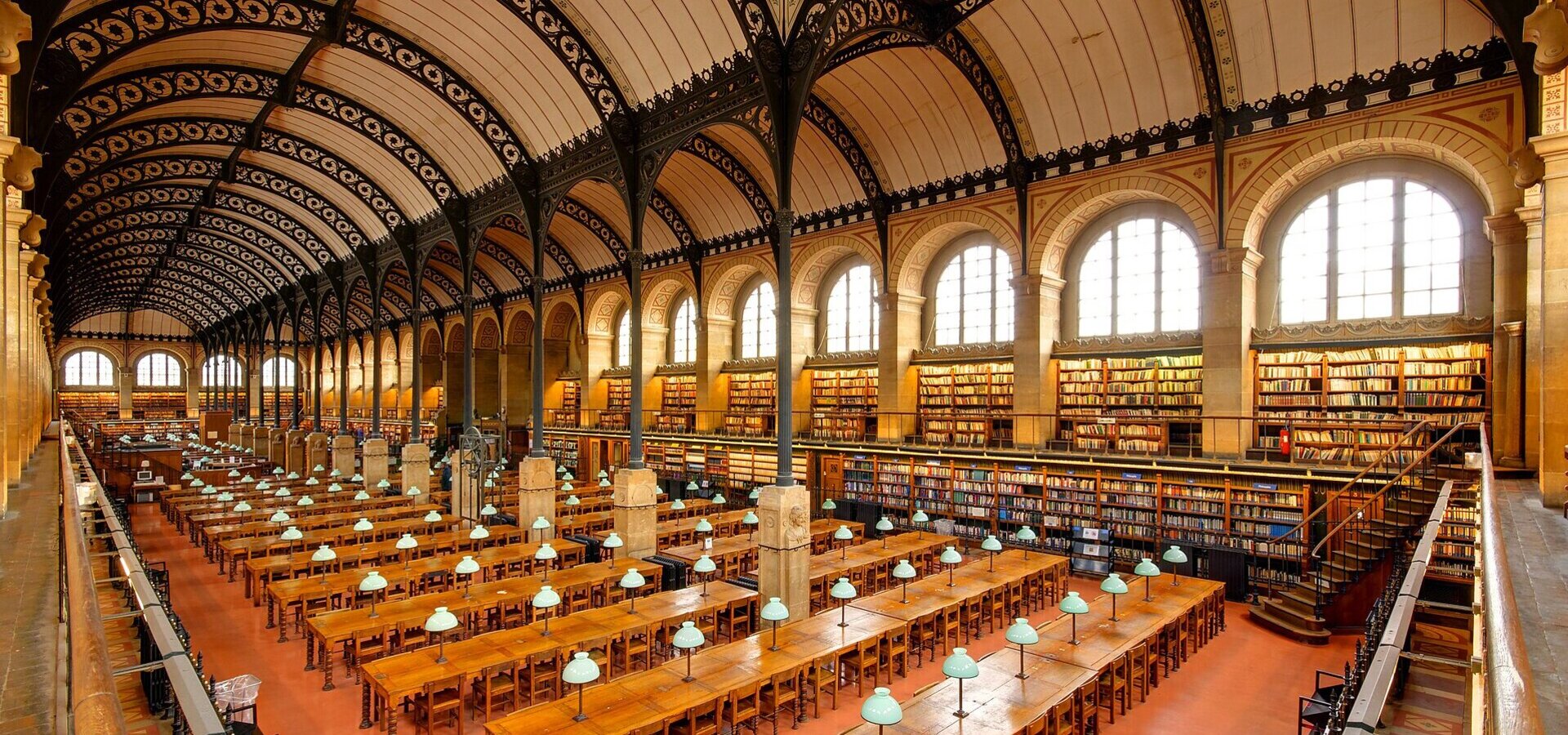 Eine große Bibliothek mit hohen Decken und historischen Holzböden. Lange Tische mit grün beleuchteten Tischlampen sind für Leser eingerichtet. Regale voller Bücher erstrecken sich entlang der Wände, und große Fenster lassen natürliches Licht herein.