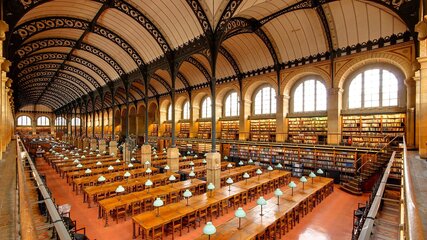 Eine große Bibliothek mit hohen Decken und historischen Holzböden. Lange Tische mit grün beleuchteten Tischlampen sind für Leser eingerichtet. Regale voller Bücher erstrecken sich entlang der Wände, und große Fenster lassen natürliches Licht herein.