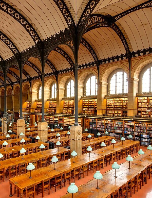 Eine große Bibliothek mit hohen Decken und historischen Holzböden. Lange Tische mit grün beleuchteten Tischlampen sind für Leser eingerichtet. Regale voller Bücher erstrecken sich entlang der Wände, und große Fenster lassen natürliches Licht herein.