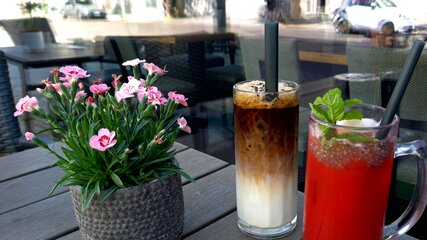 Auf dem Tisch stehen zwei Getränke: ein dunkler Eiskaffee in einem hohen Glas mit Milchschichten und ein erfrischender, roter Cocktail mit Minze. Daneben befindet sich ein Topf mit rosa Blumen.
