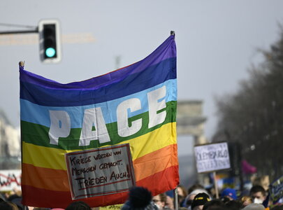 Eine Gruppe von Menschen demonstriert. Im Vordergrund wird eine große Regenbogenflagge gehalten, auf der das Wort „PACE“ in großen Buchstaben steht. Die Flagge symbolisiert Frieden und Vielfalt.
